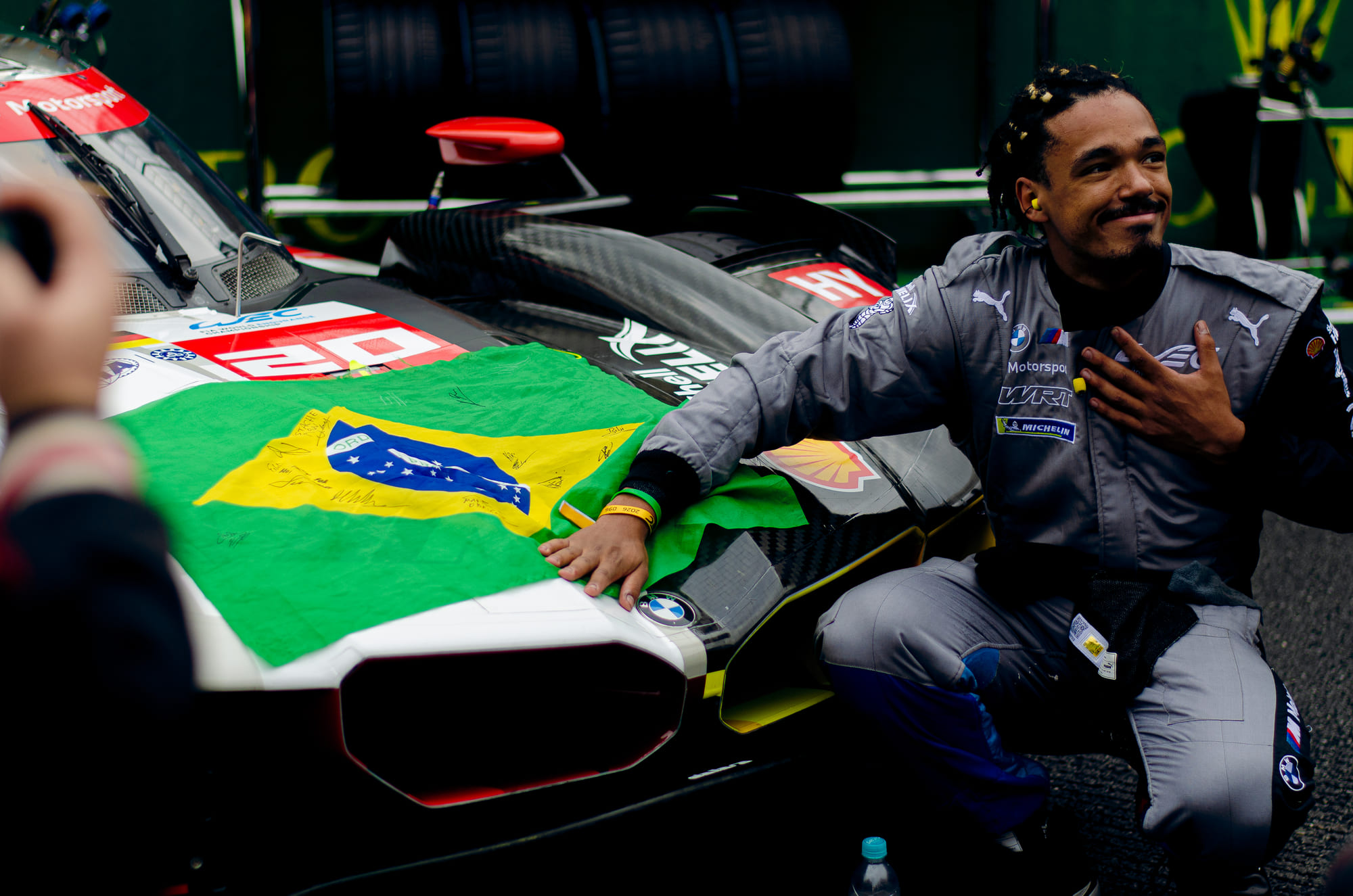 Membro da BMW M Team WRT ajoelhado ao lado do BMW M Hybrid V8 #20, da classe Hypercar, com a bandeira do Brasil autografada sobre o bico durante o grid walk no WEC 6 Horas de São Paulo 2026, em Interlagos