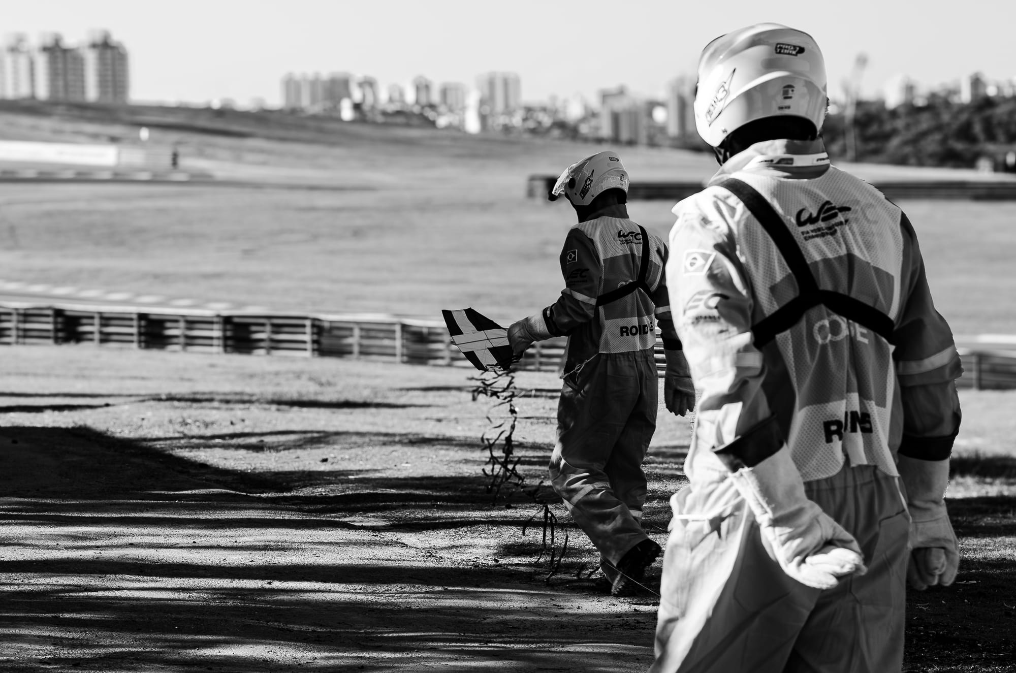 Dois fiscais do WEC recolhendo destroços na área de escape, com a cidade ao fundo, em preto e branco, no WEC 6 Horas de São Paulo 2026, em Interlagos